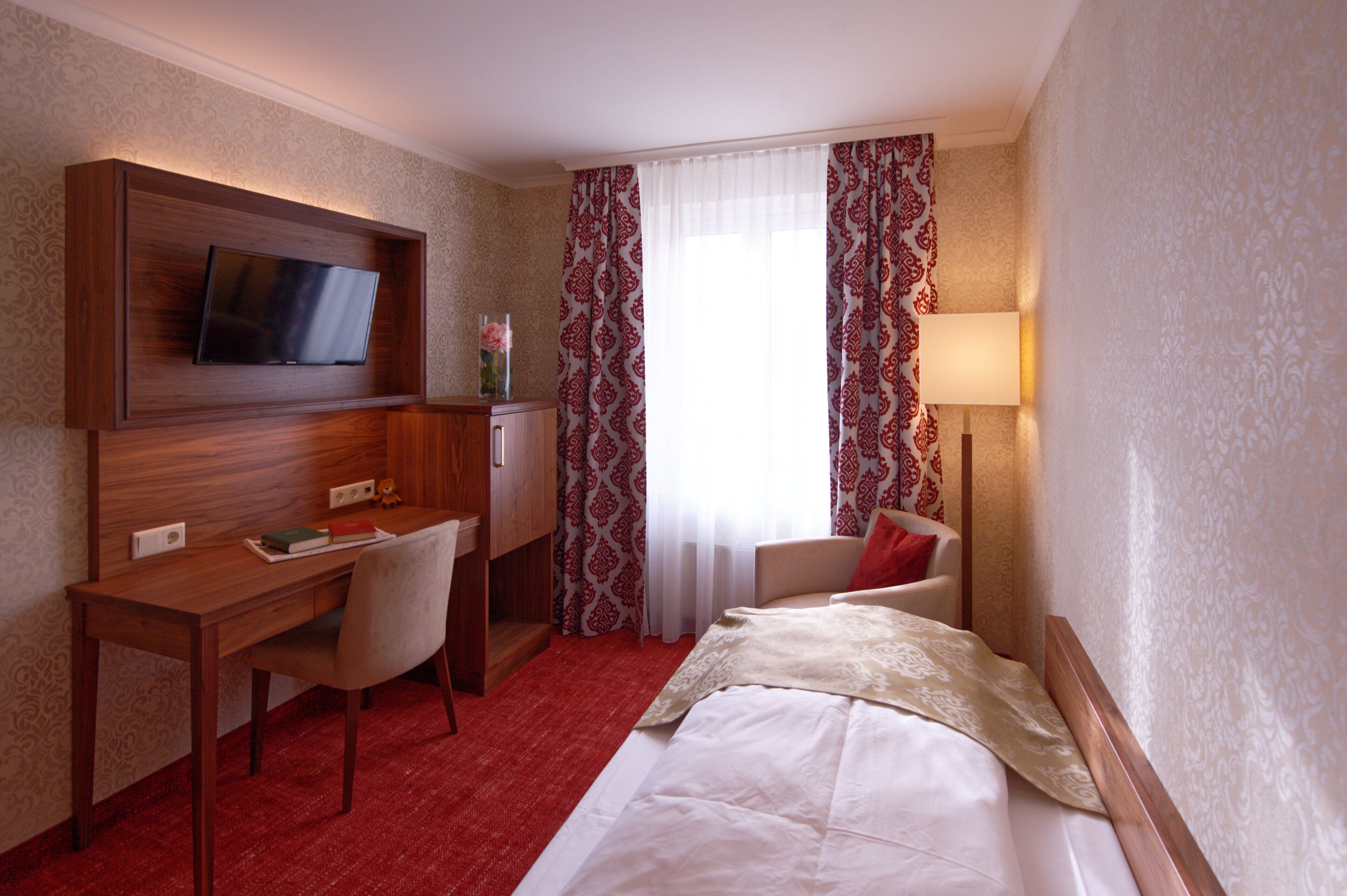 A hotel room with a bed, a desk with a chair, a television mounted on the wall, and a window with patterned curtains.