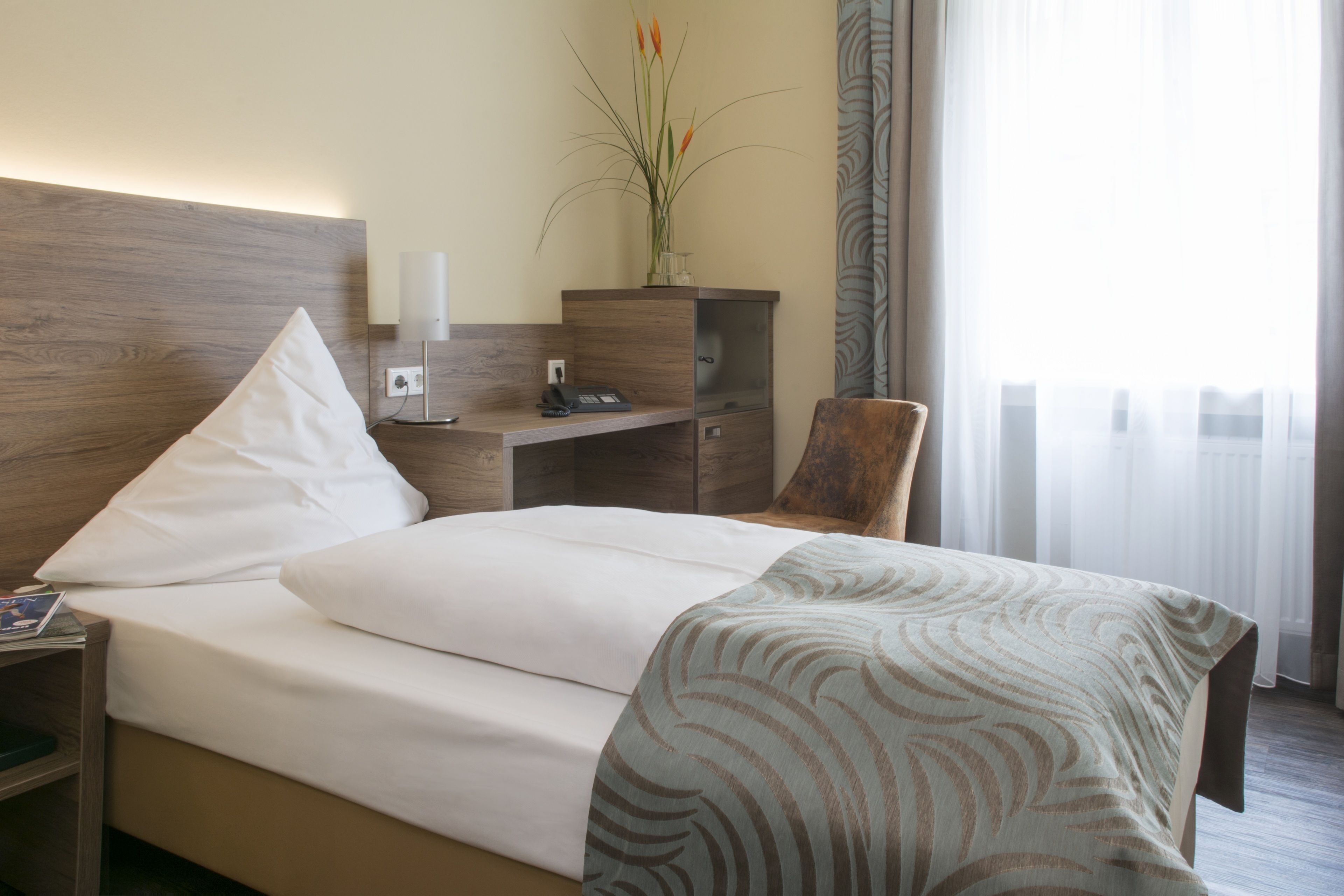 Una habitación de hotel con cama, escritorio, silla, mesita de noche y un jarrón con flores.