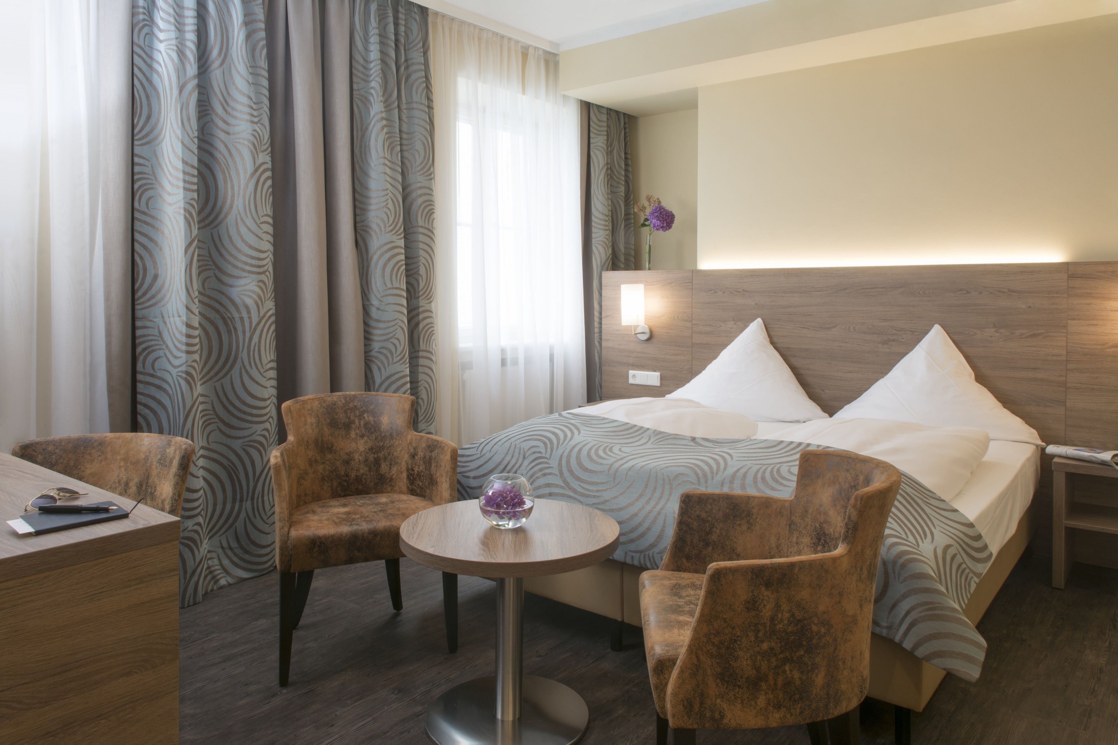 A hotel room with a bed, two armchairs, a small round table, and a desk with a chair.