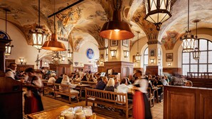 A crowded beer hall with wooden benches and tables, large beer mugs, and ornate ceiling decorations. - Hotel Schlicker (Munich)