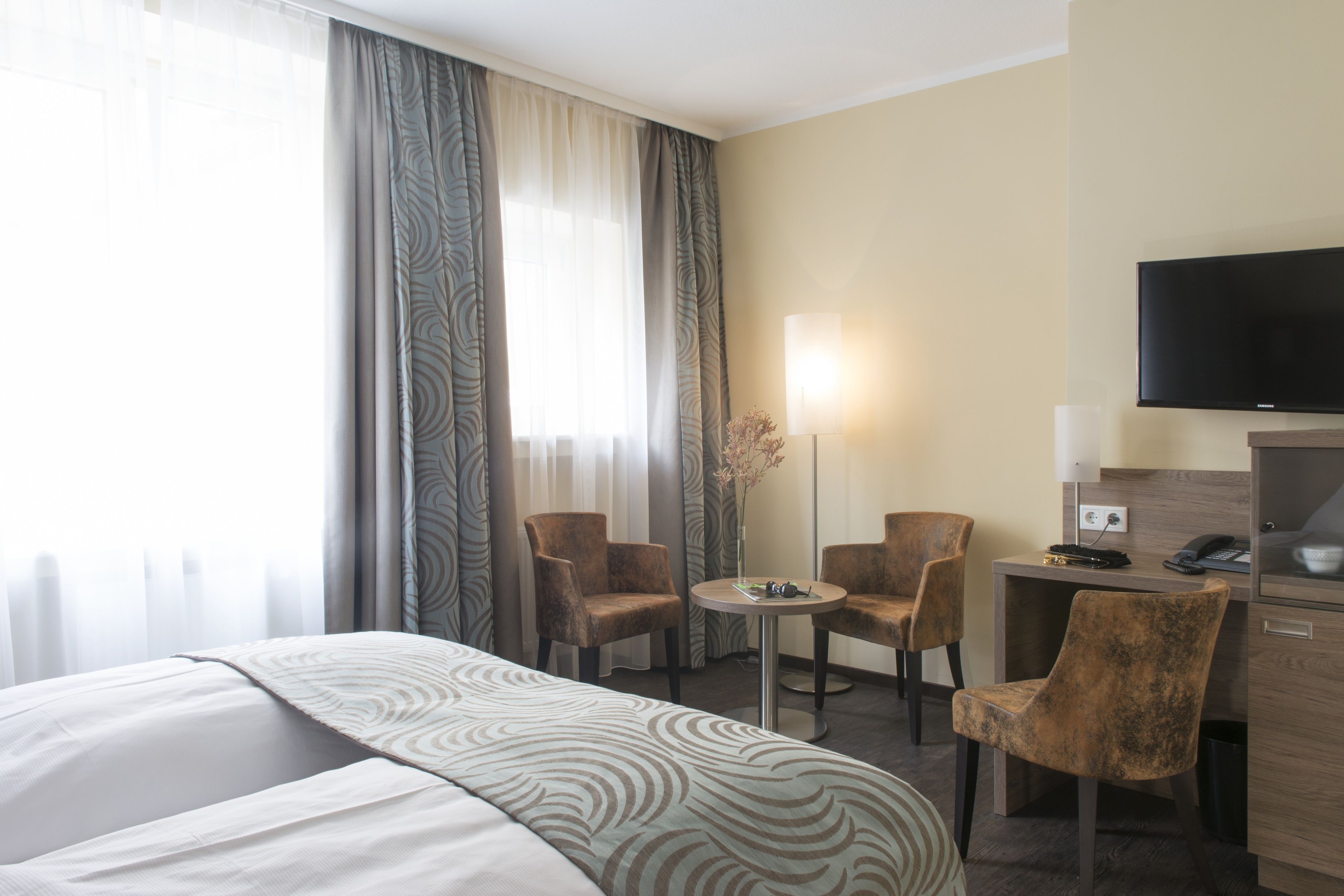 Ein Hotelzimmer mit einem Bett, zwei Sesseln, einem Schreibtisch, einem Flachbildfernseher und einer Lampe.
