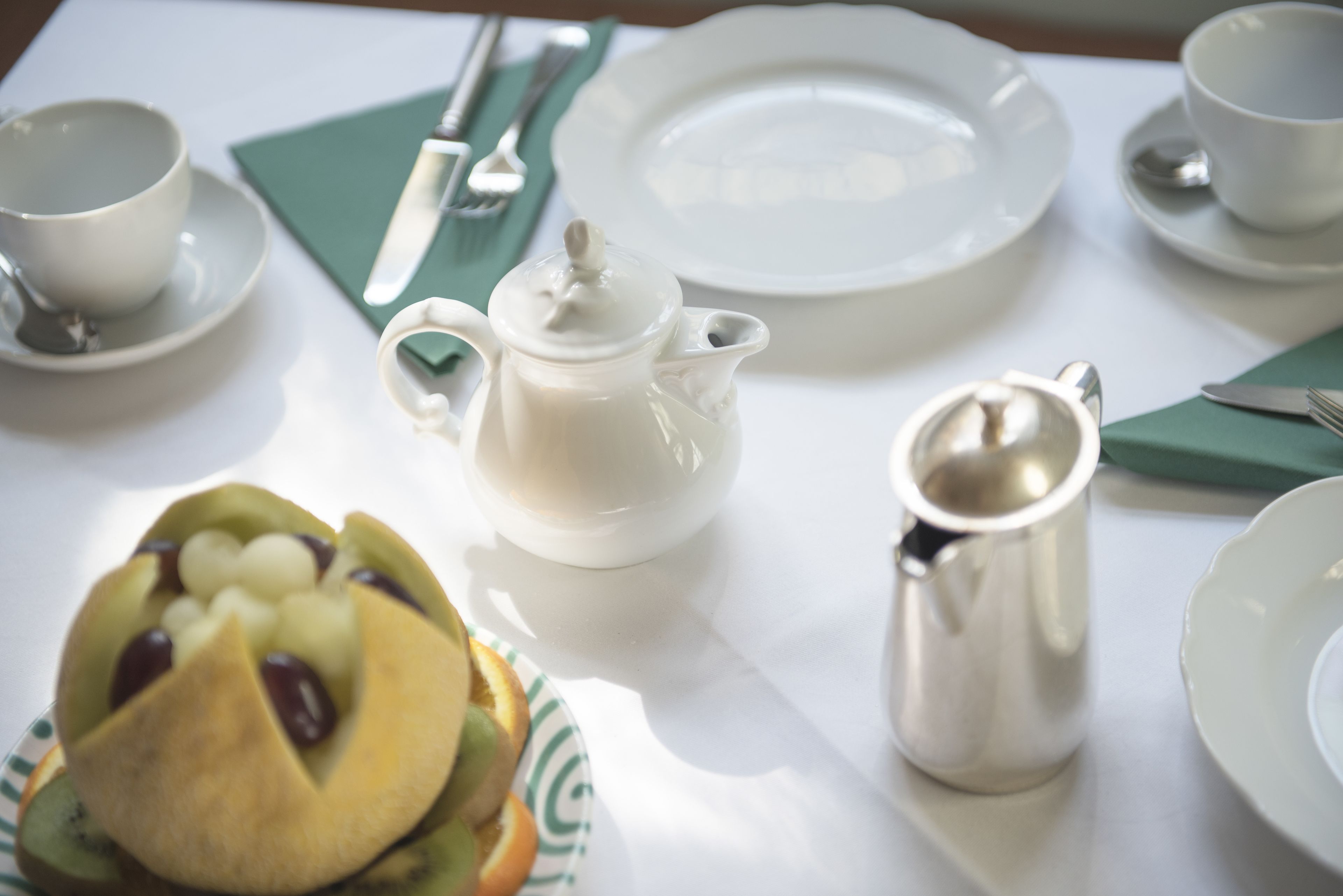 A table set with white plates, a teapot, a cup, a silver pitcher, and a fruit arrangement.