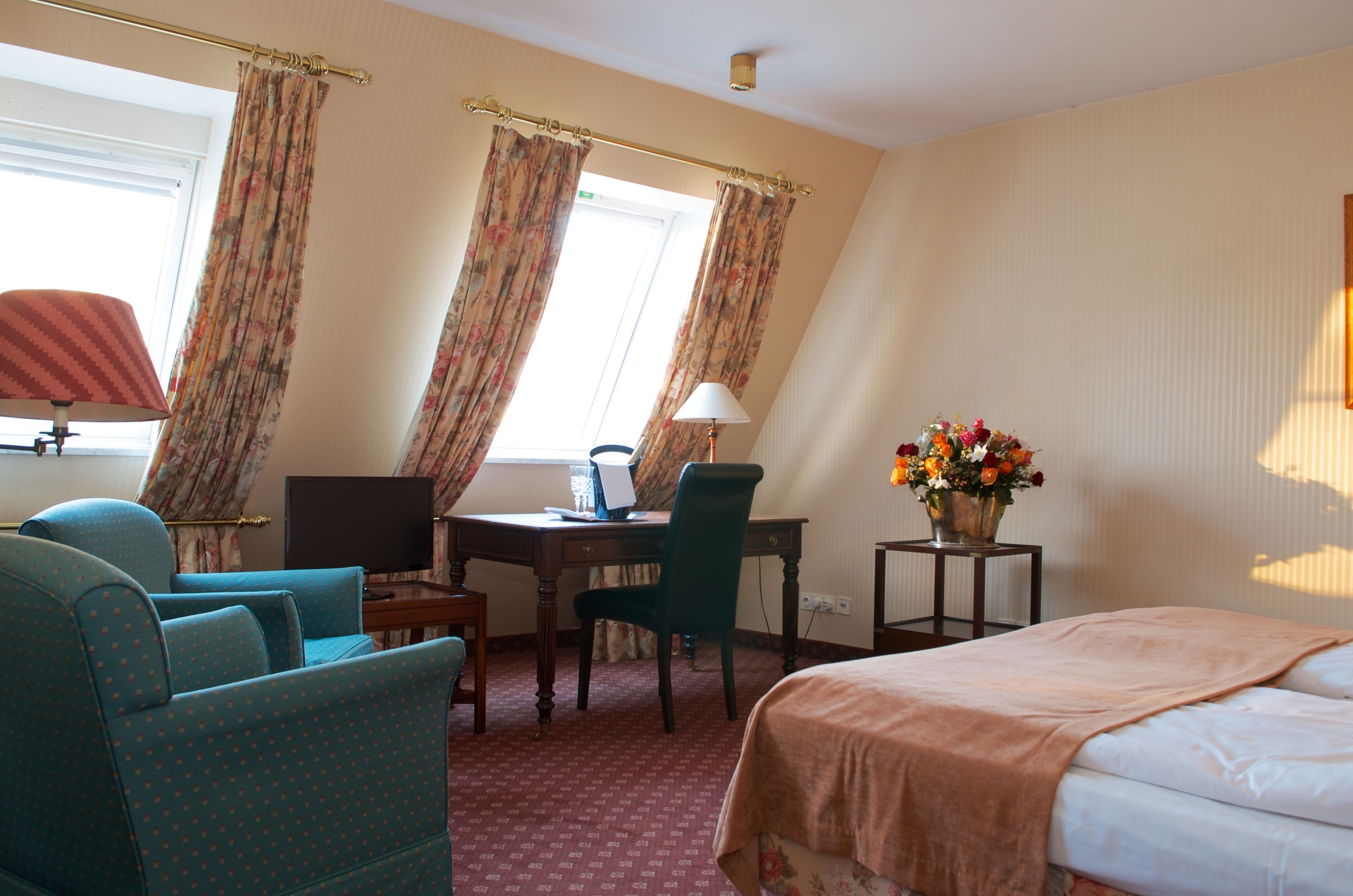 A hotel room with a bed, a desk with a lamp, a chair, a TV, and a vase with flowers.