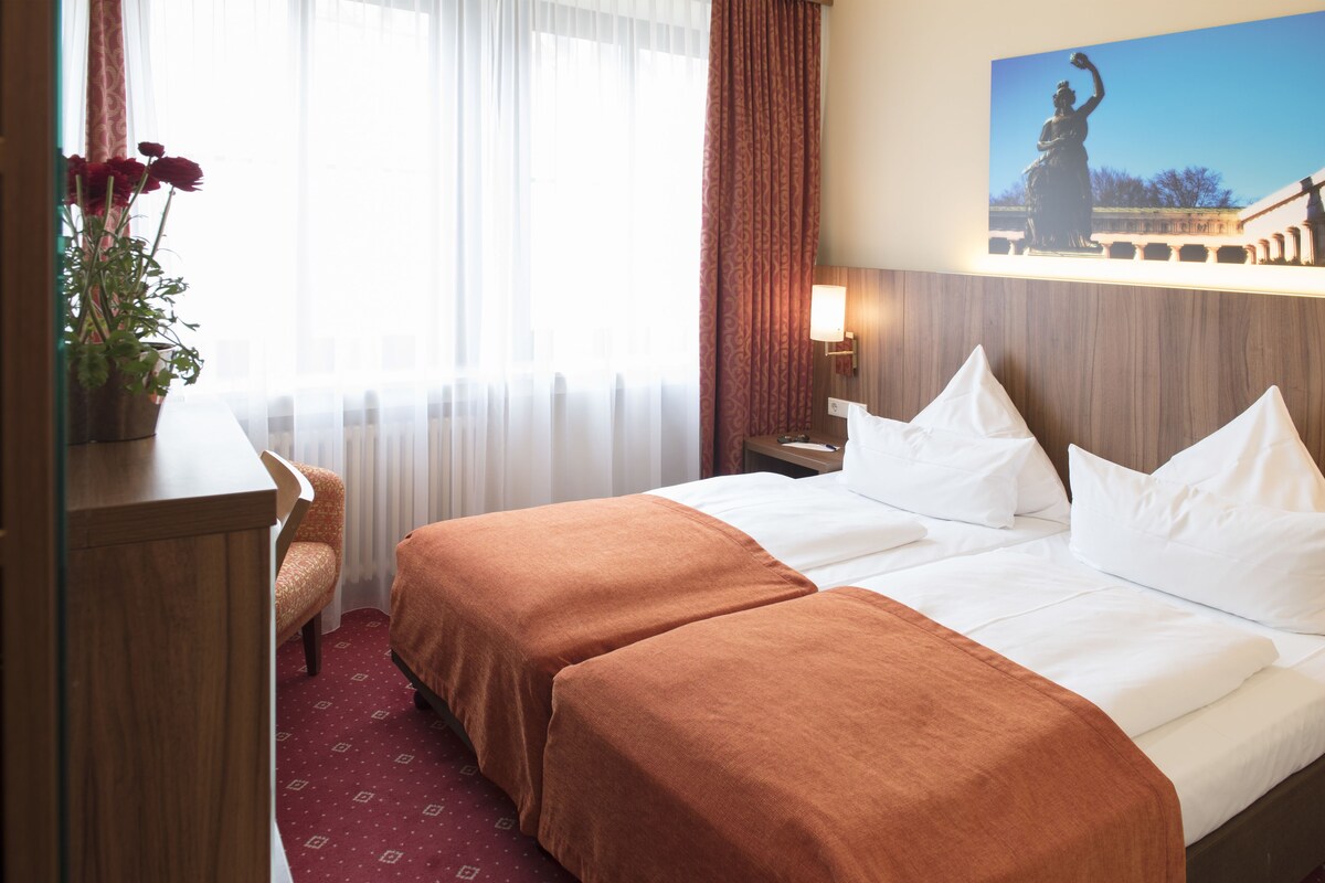 A hotel room with two beds, a desk, a chair, a potted plant, and a painting on the wall.