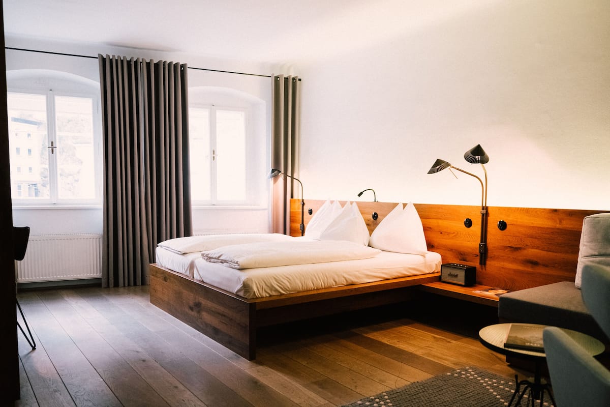 Un dormitorio con cabecera de madera, cama con ropa blanca, mesita de noche con lámpara, una silla y vista a la ciudad a través de una ventana con cortinas.