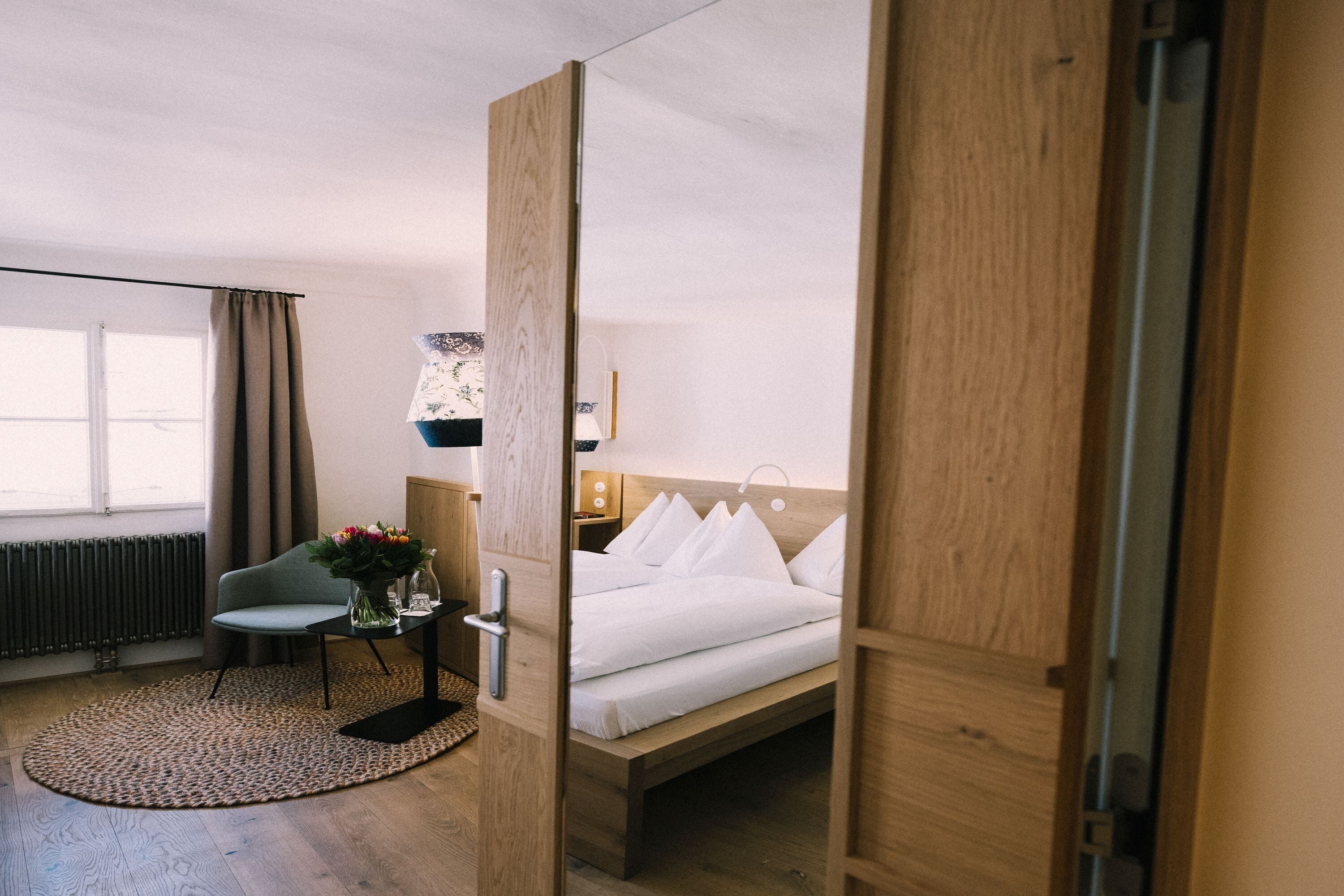 A hotel room with a bed, a chair, a small table with a vase of flowers, a lamp, and a view of the outdoors through a window.