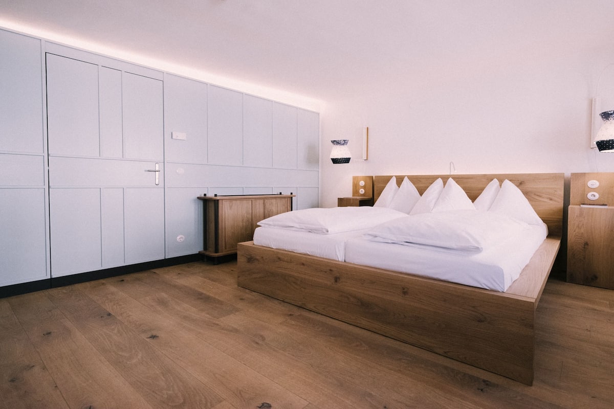 Un dormitorio con cama de madera, ropa de cama blanca, una mesita de noche de madera y un armario con acabado en tonos claros.