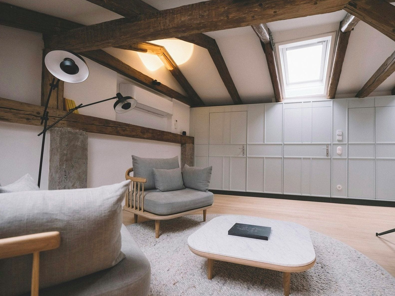 A loft-style room with exposed wooden beams, a built-in wardrobe, a cushioned seating area, and a floor lamp.