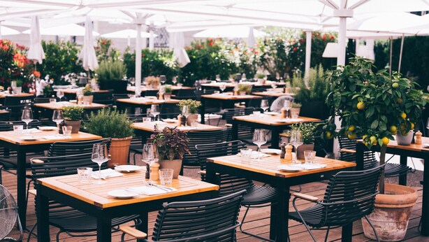 Un restaurante al aire libre con varias mesas y sillas, rodeado de macetas con plantas y sombrillas.