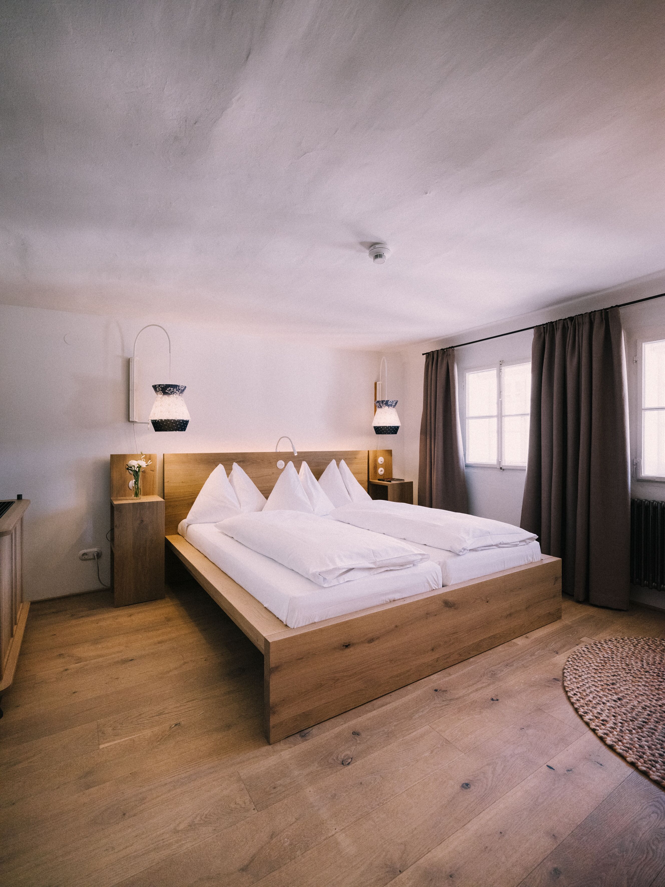 A bedroom with a wooden bed, two bedside tables, a wall-mounted lamp, and a window with curtains.