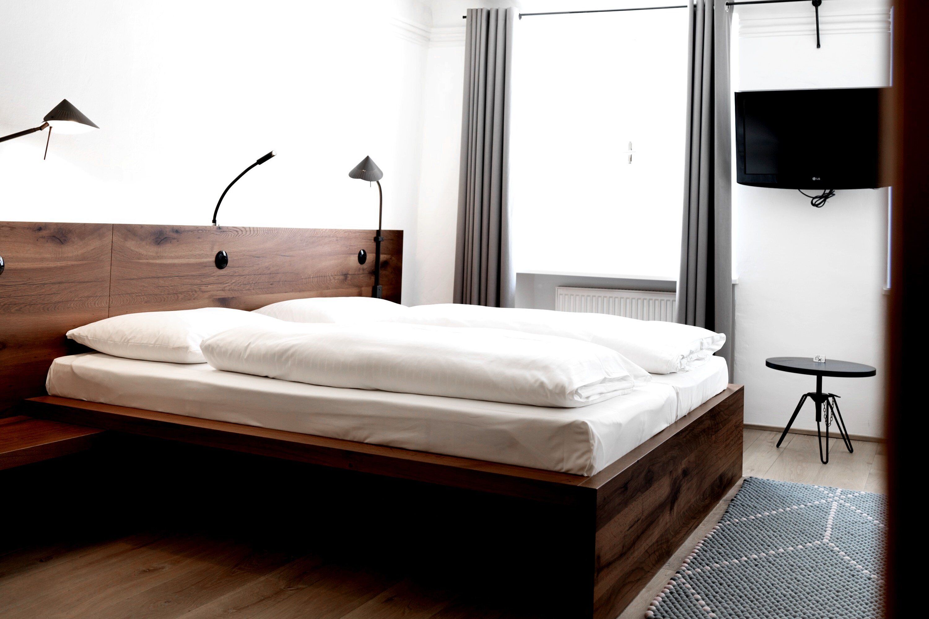Ein modernes Schlafzimmer mit einem Holzkopfteil, einem ordentlich bezogenen Bett, einem kleinen runden Tisch und einem an der Wand montierten Fernseher.