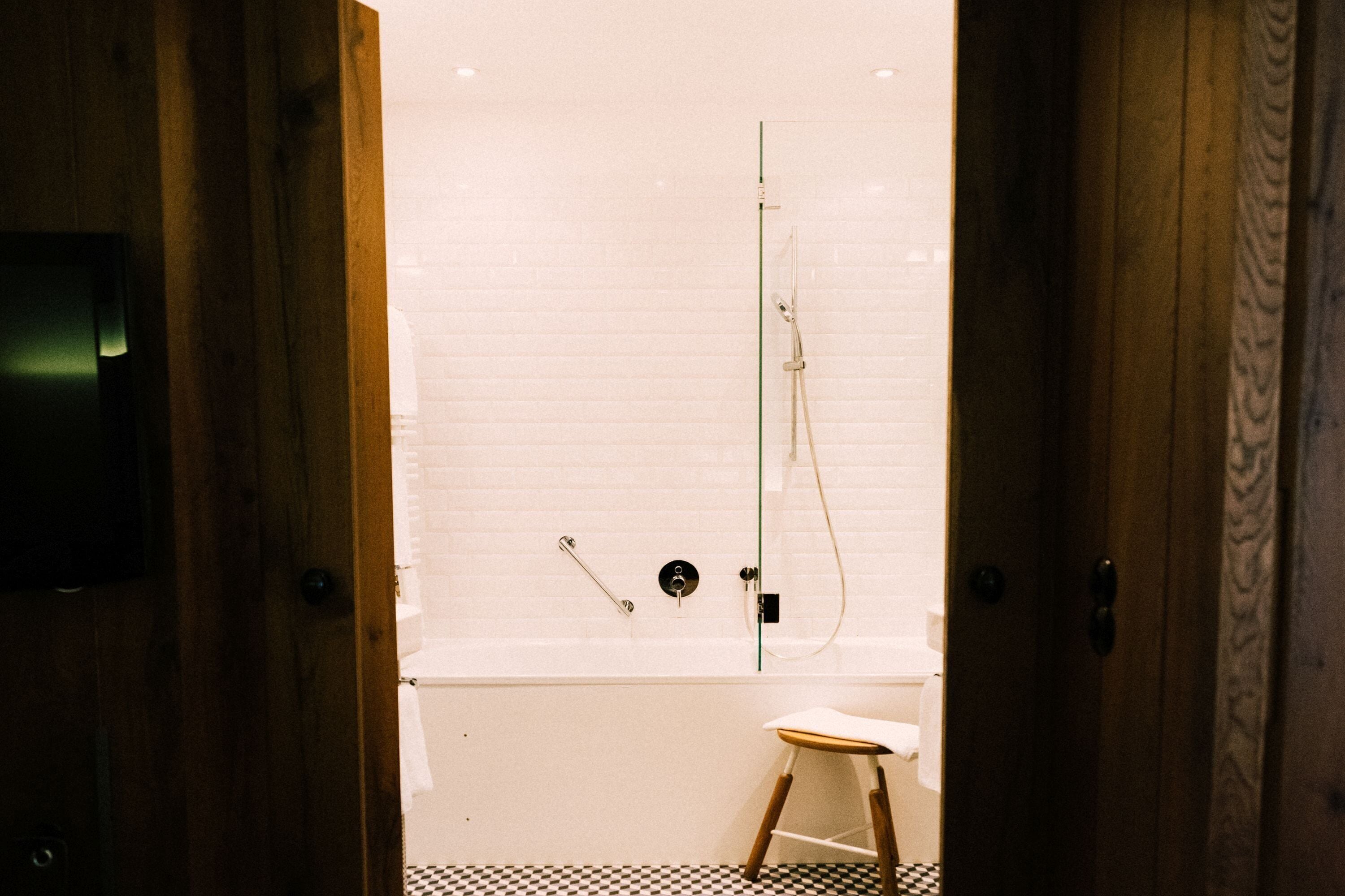 A bathroom with a white tiled shower, a wooden stool, and a checkered floor.