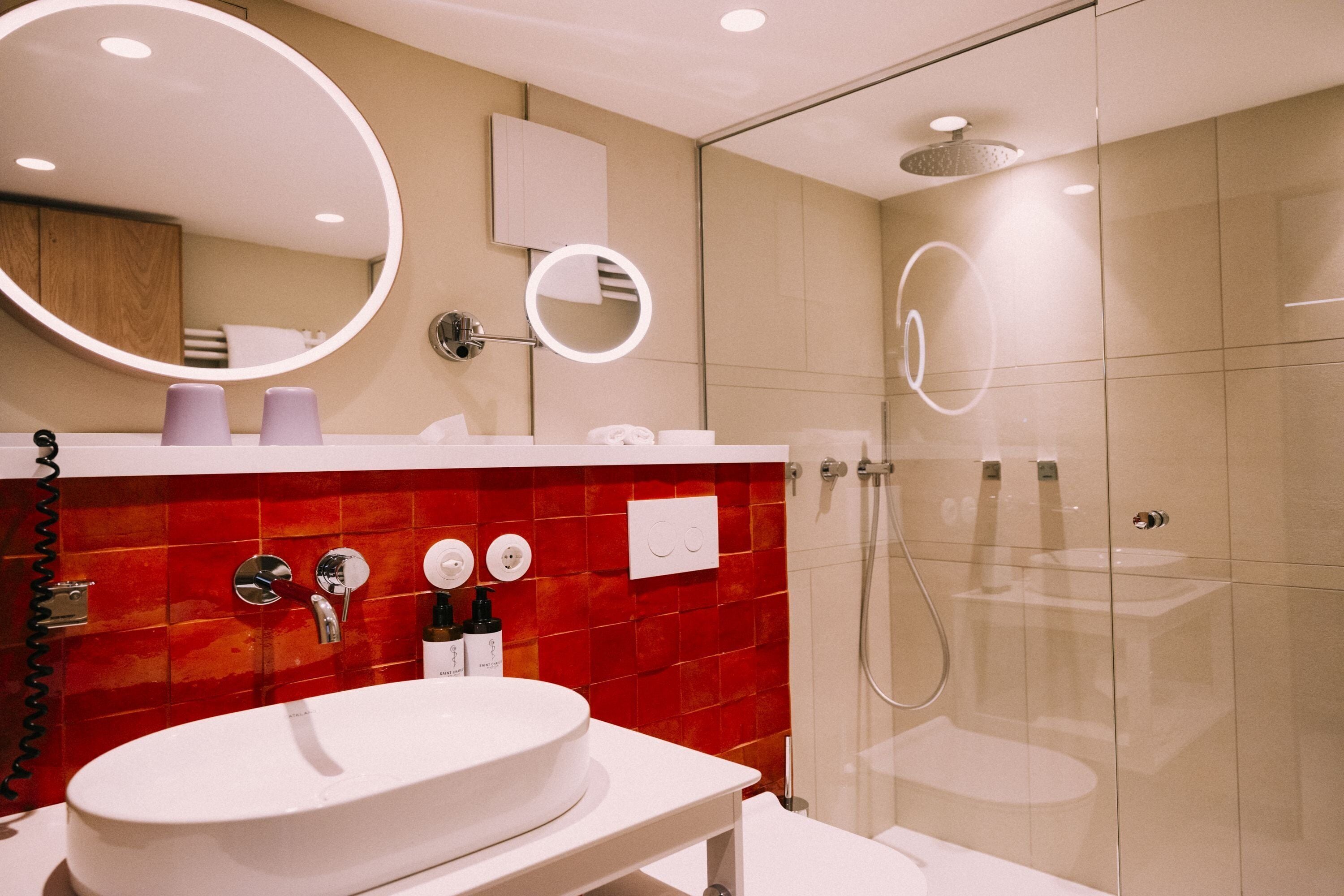 Un baño moderno con lavamanos blanco, salpicadero de azulejos rojos, dos espejos y ducha con mampara de vidrio.
