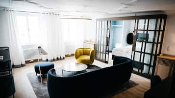 Una sala moderna con un sofá, un sillón amarillo y una mesa de centro redonda. Se observa una ventana grande con cortinas, un radiador y una puerta que da a otra habitación.