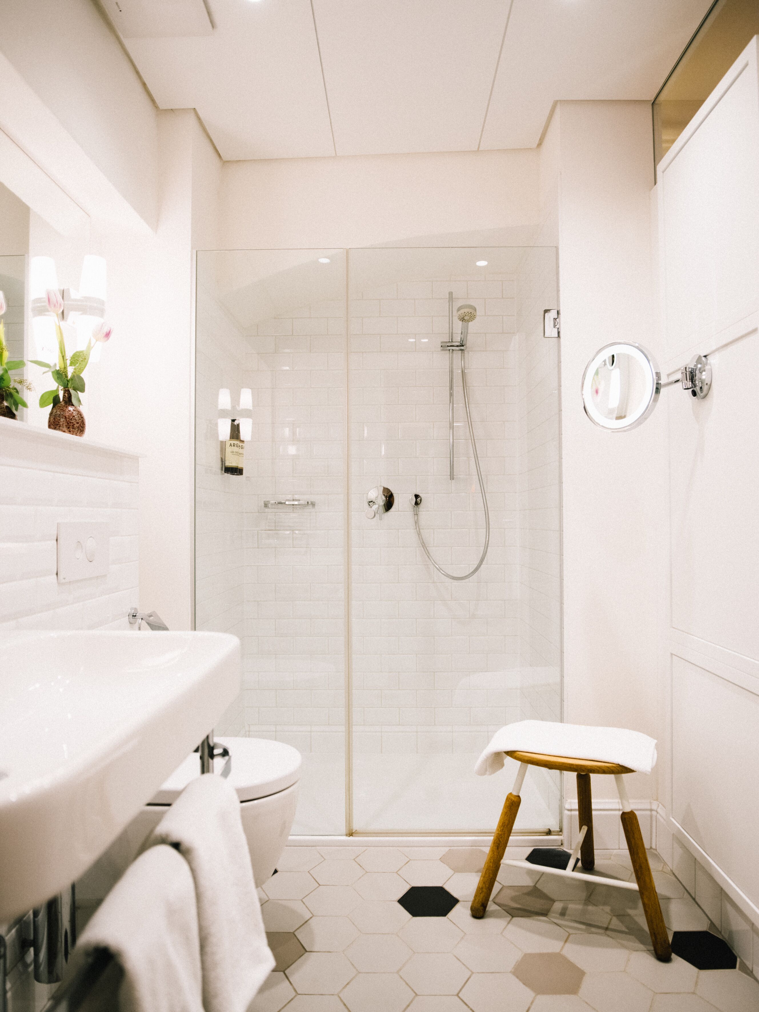 Un baño moderno con ducha de vidrio, lavamanos blanco y un taburete de madera pequeño.