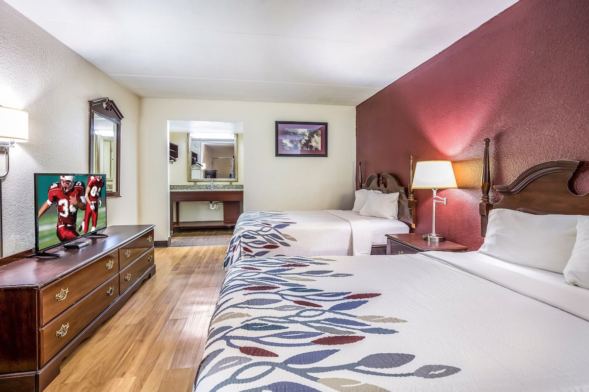 Habitación de hotel con dos camas, cómoda, televisor, espejo, mesita de noche con lámpara y un cuadro en la pared.