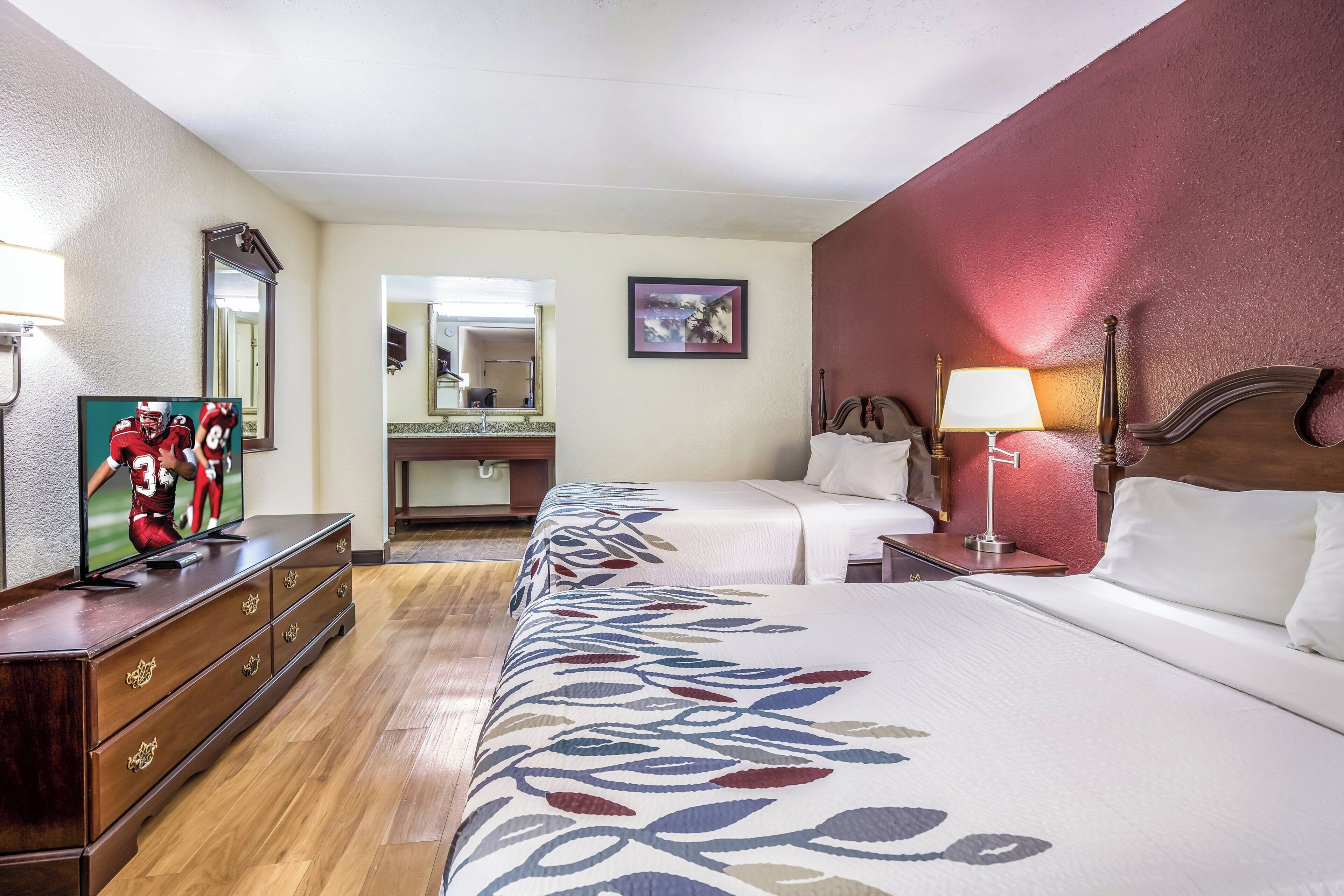 Une chambre d’hôtel avec un grand lit, une commode, une télévision, un miroir, une table de chevet avec une lampe et un tableau au mur.