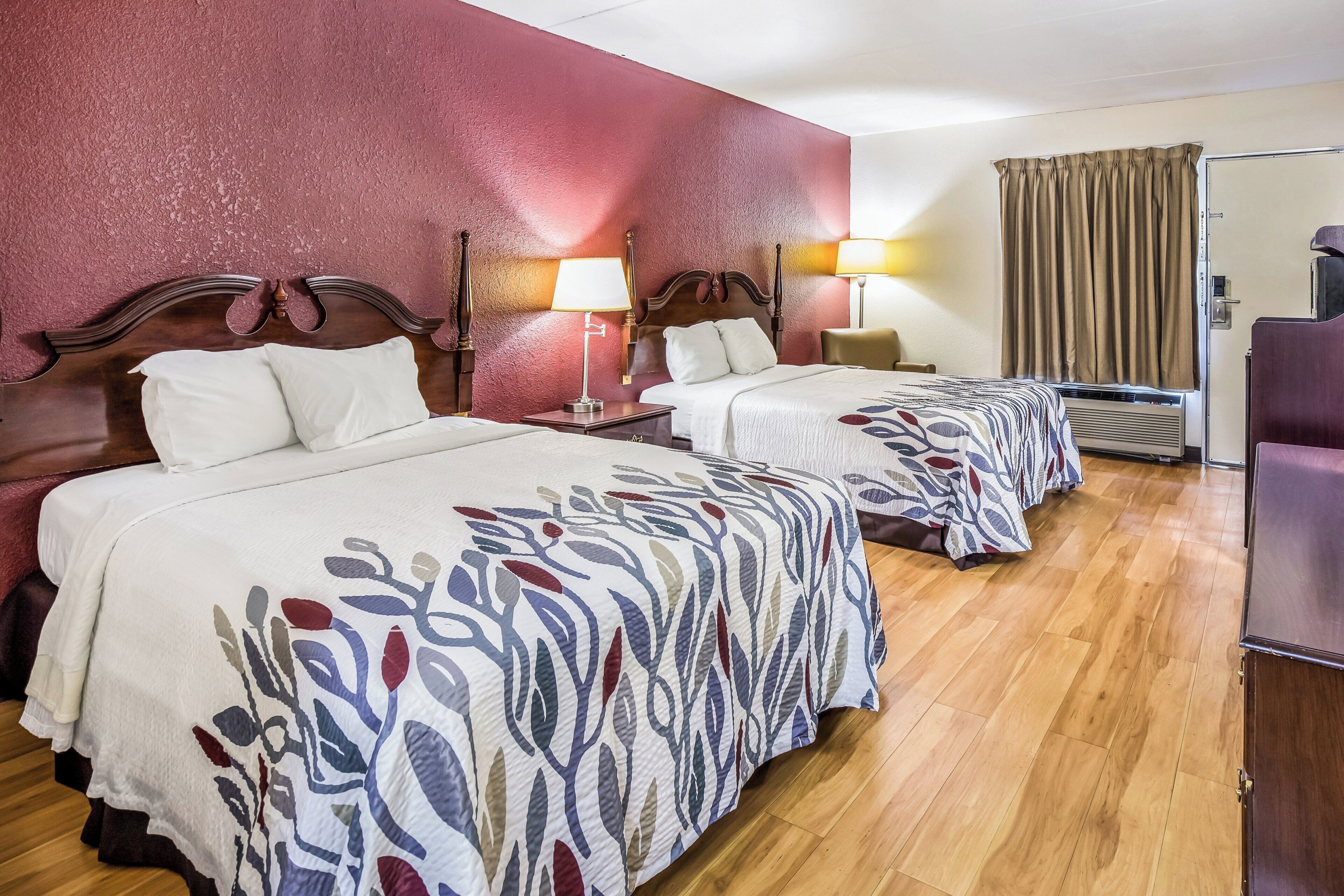 Habitación de hotel con dos camas, piso de madera, pared de acento roja y una ventana con cortinas.