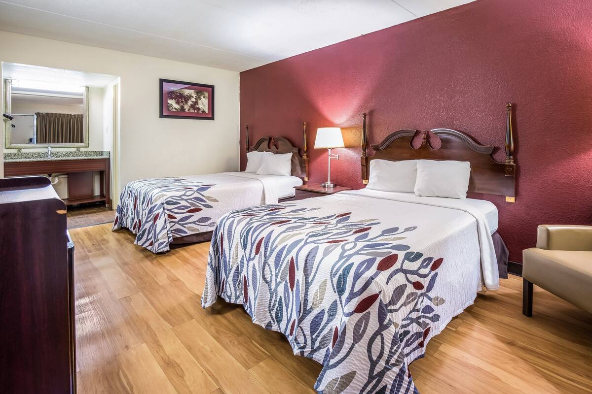 Habitación de hotel con dos camas, suelo de madera, pared decorada en rojo, un espejo y un escritorio pequeño.