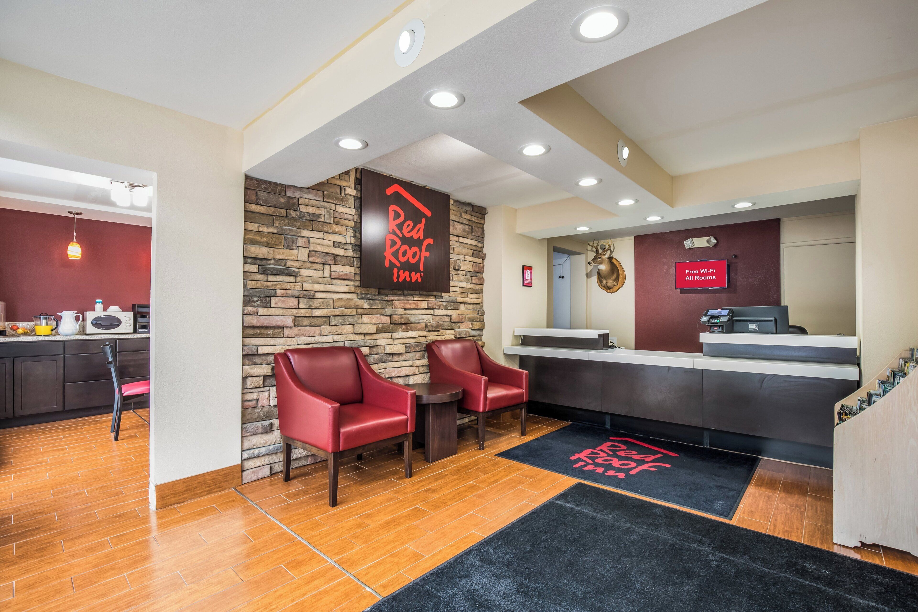 Un espace d’accueil d’un hôtel Red Roof Inn, avec un mur en pierre, deux chaises rouges, un comptoir d’accueil et un tapis noir et rouge.