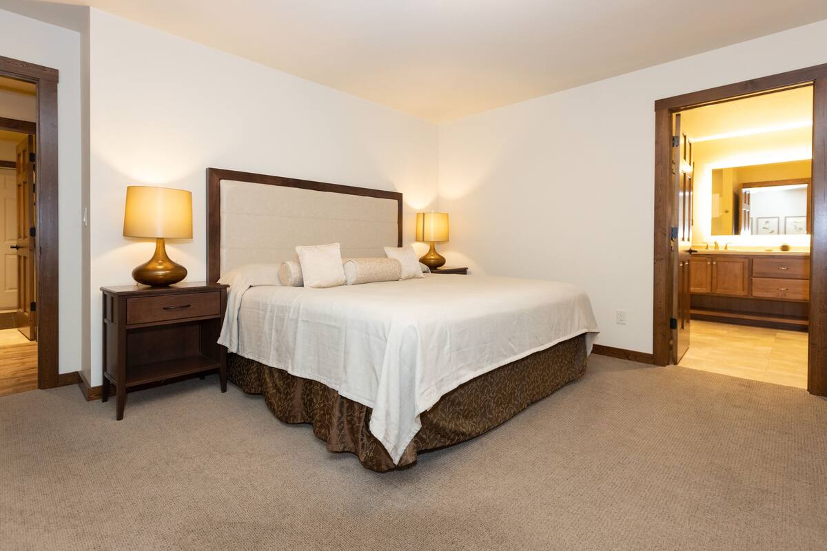 A neatly arranged bedroom with a large bed, two bedside tables with lamps, a wooden headboard, and a view into the bathroom through an open door.