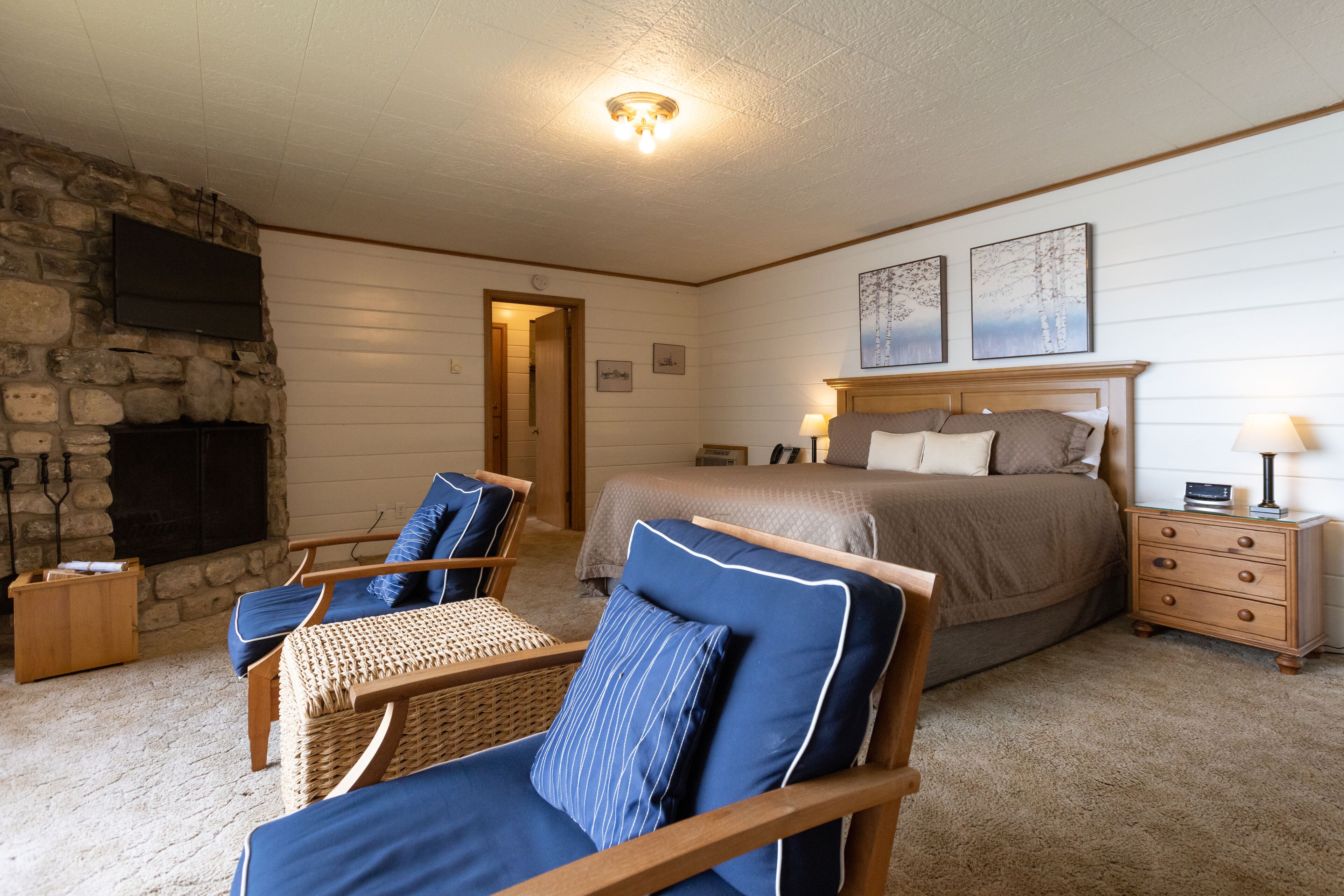 Une chambre avec une cheminée en pierre, un lit avec une tête de lit en bois, deux téléviseurs fixés au mur et un fauteuil en osier avec des coussins bleus.