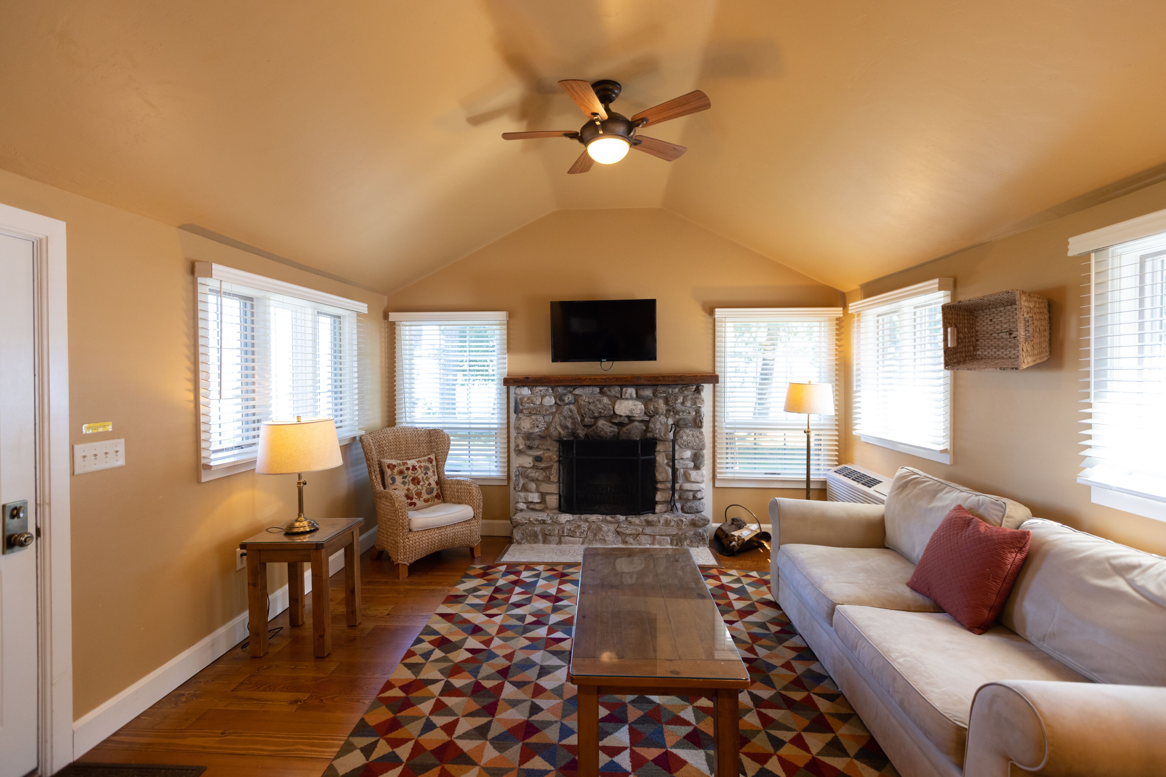 A cozy living room with a fireplace, a ceiling fan, a sofa, a coffee table, and a patterned rug.