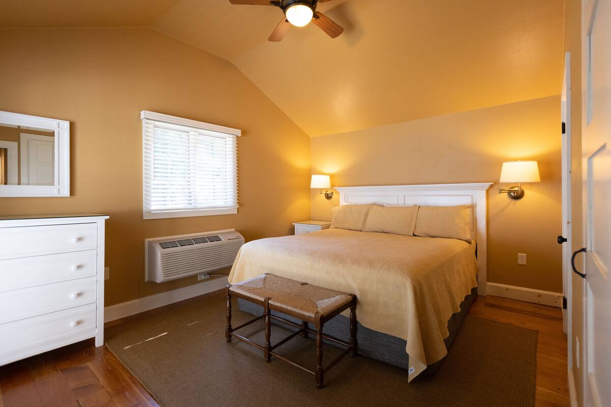 A bedroom with a bed, bedside lamps, a ceiling fan, a window with blinds, a dresser, and a bench.