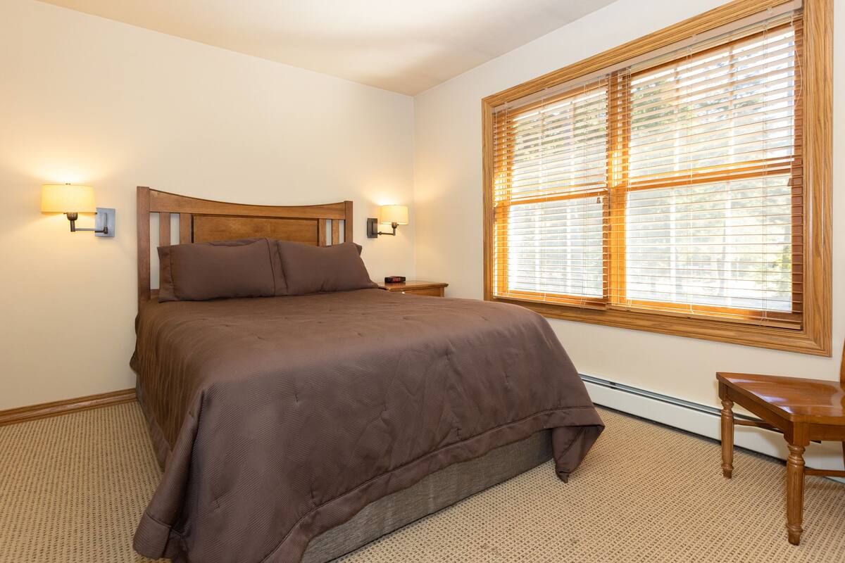 A bedroom with a large bed, two bedside lamps, a wooden nightstand, and a window with blinds.