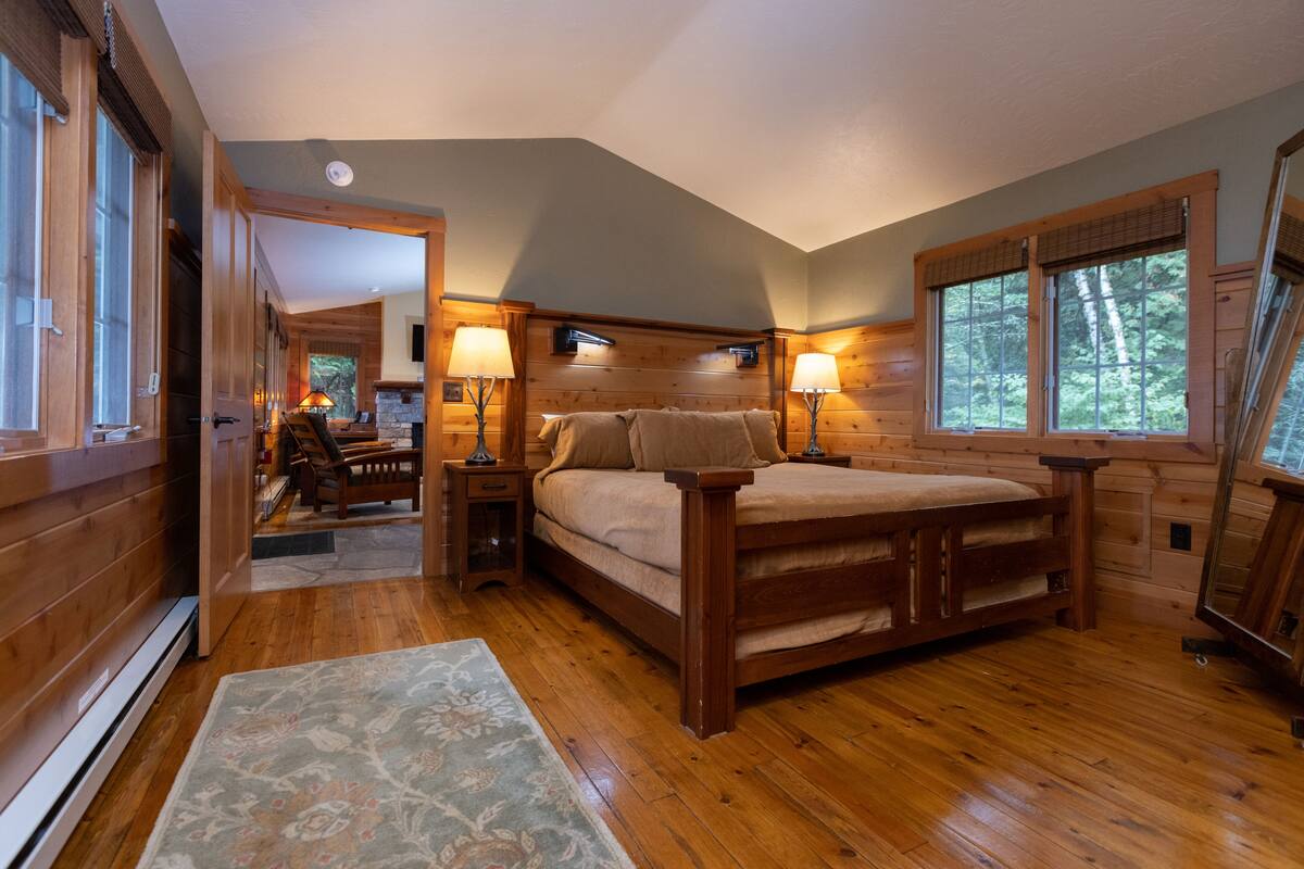 A wooden cabin bedroom with a large bed, two bedside lamps, and a view of the forest through the windows.