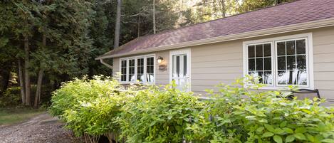 A house with a wooden deck, surrounded by dense trees and a gravel path.