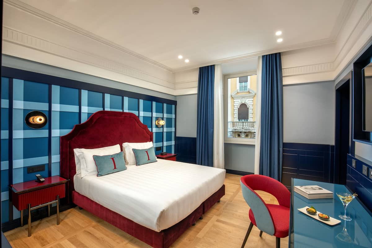 A hotel room with a large bed, a red headboard, a nightstand, a red chair, a glass table with a martini glass, and a view of a building through a window.
