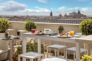 A rooftop terrace with tables set for breakfast, including croissants, fruit, and orange juice, overlooking a cityscape with historic buildings. - The Independent Hotel (Rome)