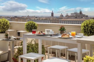 A rooftop terrace with tables set for breakfast, including croissants, fruit, and orange juice, overlooking a cityscape with historic buildings.