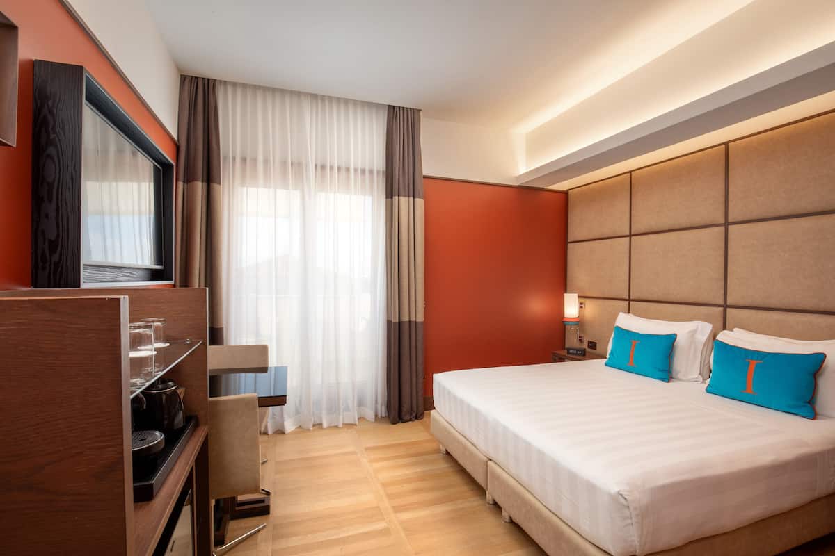 A hotel room with a large bed, a TV, a desk with a chair, and a window with curtains.