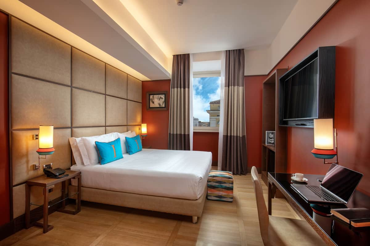 A hotel room with a large bed, a desk with a laptop, a TV, and a window with curtains.