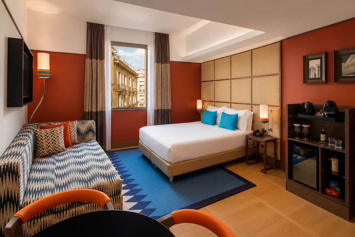 A hotel room with a bed, a sofa, a desk, a TV, and a view of buildings outside the window.