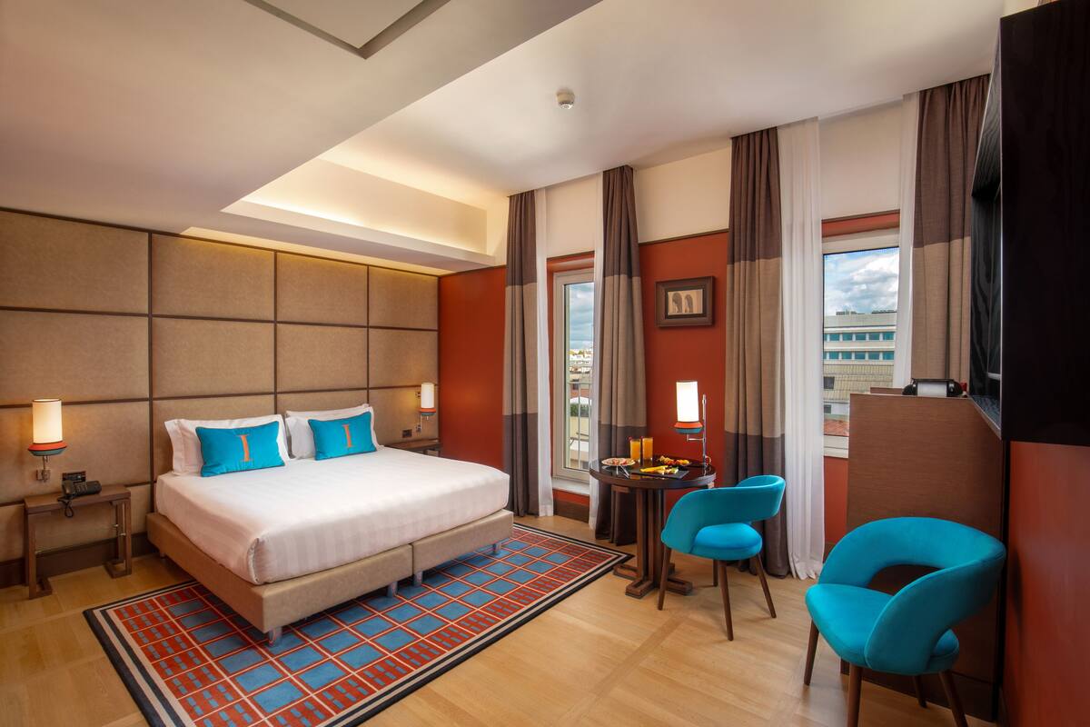 A hotel room with a large bed, two bedside tables, a desk with a chair, a view of the city, and a window with curtains.