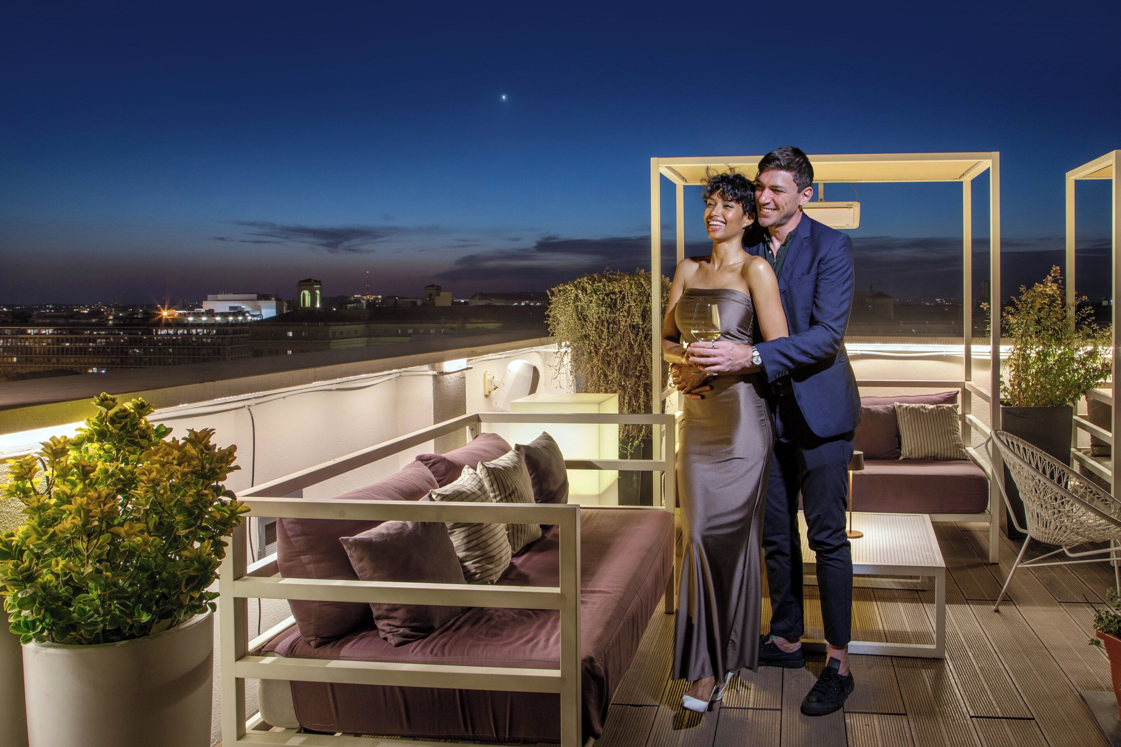 A couple on a rooftop terrace with a city view, a sofa, and a table.