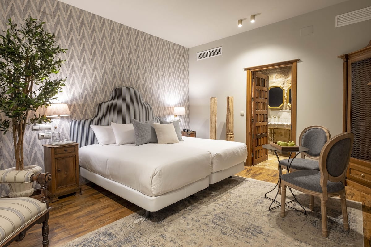 A bedroom with a large bed, bedside tables, a chair, a nightstand, a lamp, a mirror, and a door leading to another room.
