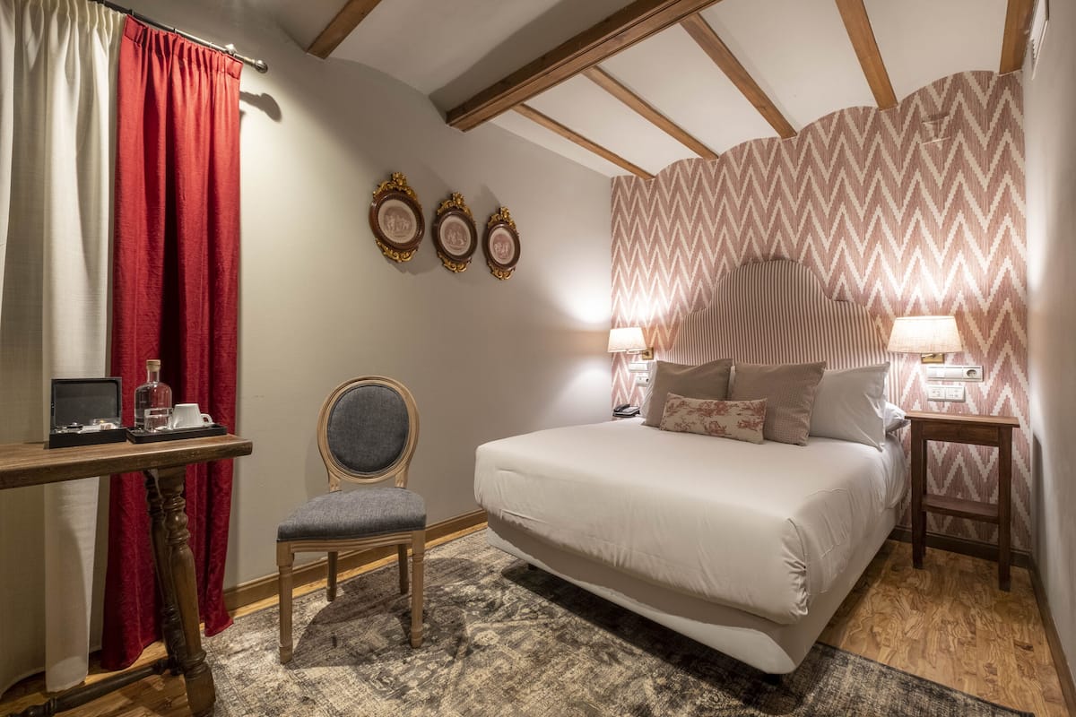 A bedroom with a large bed, a desk with a chair, a lamp, and a wall with three framed pictures.