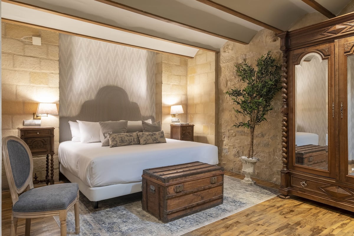 A bedroom with a large bed, bedside lamps, a wooden chest, a chair, a mirror, and a potted plant.