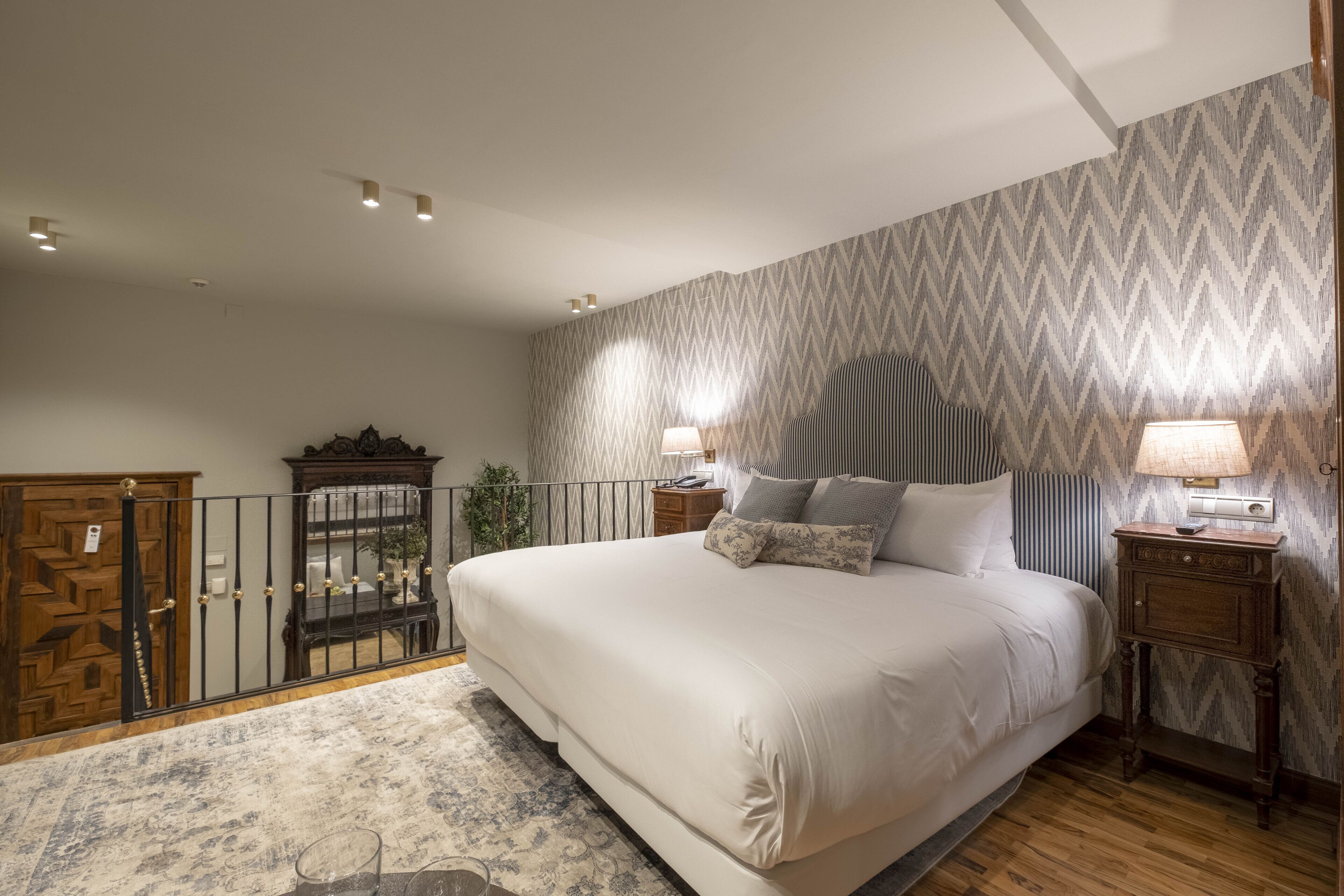 A bedroom with a large bed, two bedside tables with lamps, a wooden wardrobe, and a patterned wallpaper.