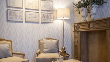 A cozy living room with a fireplace, two armchairs, a coffee table, and framed architectural drawings on the wall.