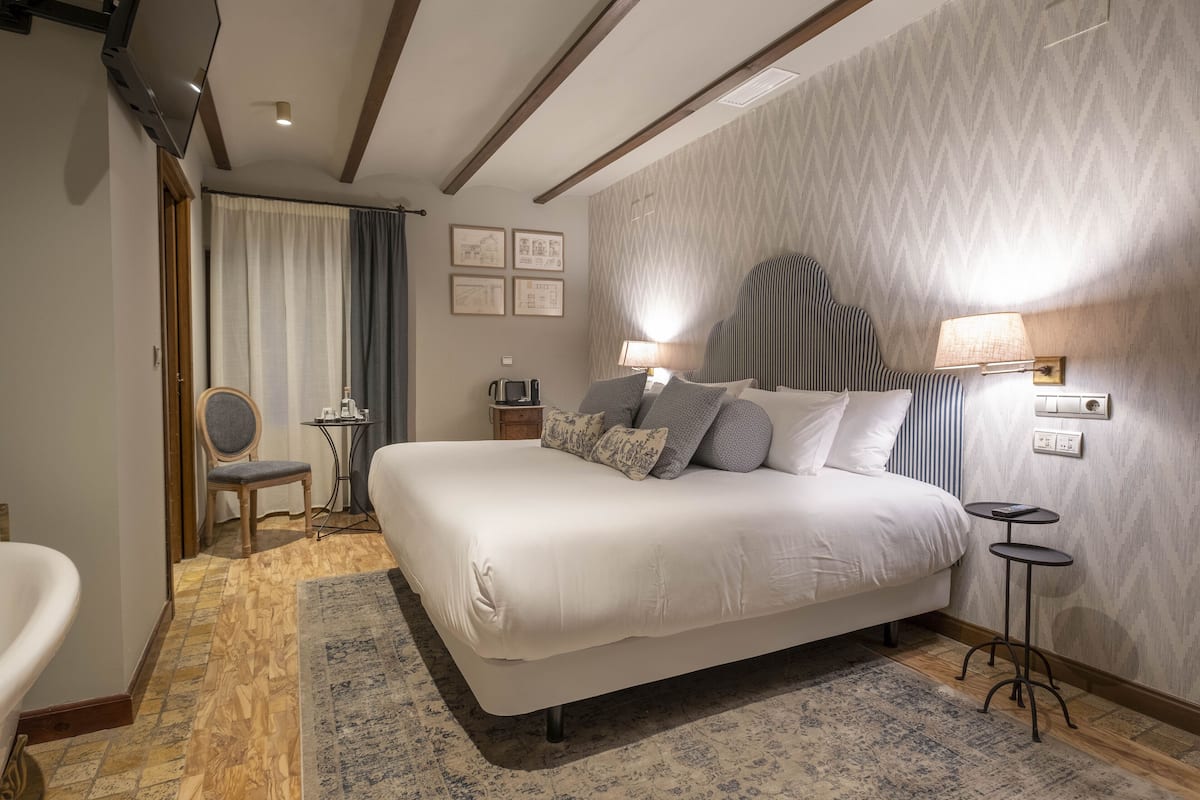 A modern hotel room with a large bed, a small side table, a chair, and a desk with a lamp.