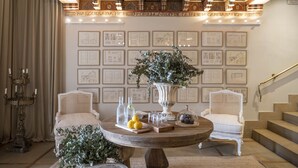 A room with a round wooden table, two white upholstered chairs, a large vase with greenery, and framed architectural drawings on the wall.