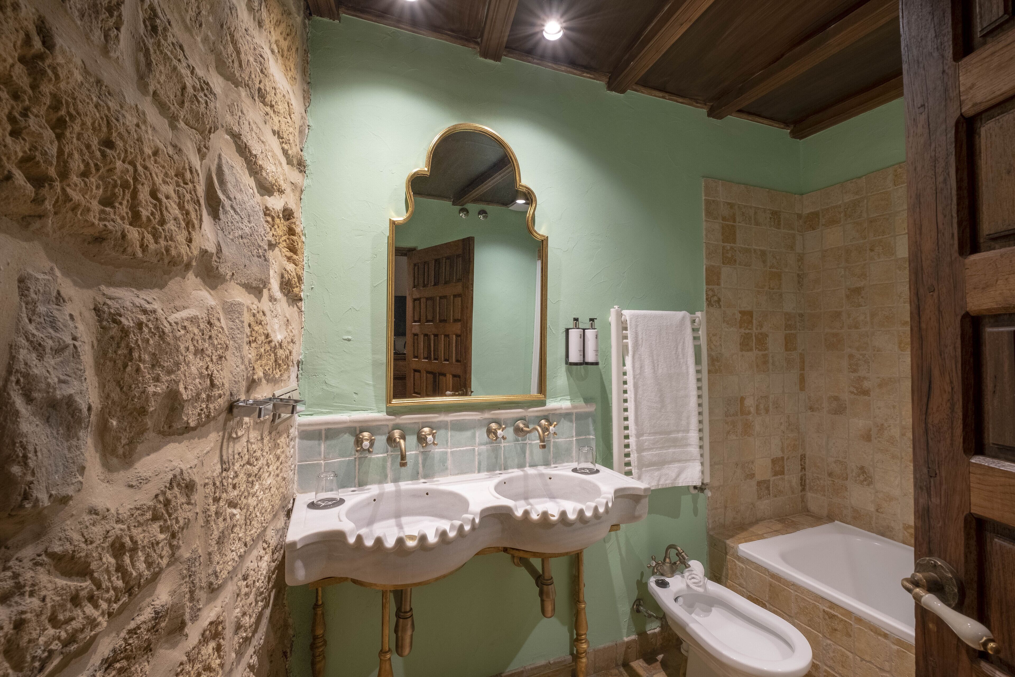 Une salle de bain avec un mur en pierre, une porte en bois, un miroir doté d’un cadre orné et une baignoire.