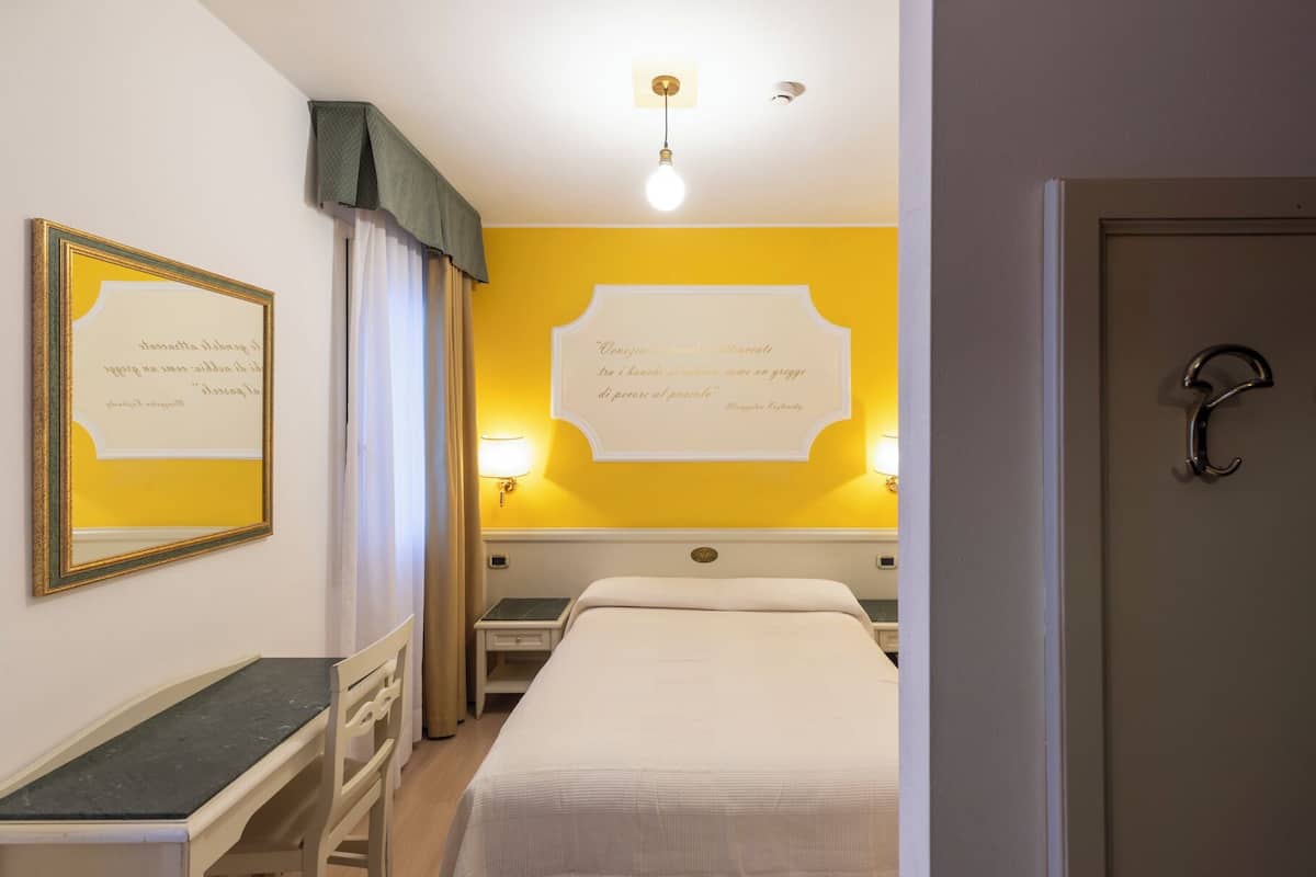 A hotel room with a bed, a desk, and a chair. There are two wall-mounted lamps and a framed quote on the wall.