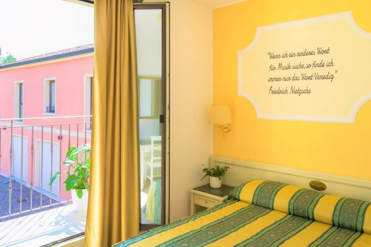 A hotel room with a bed, a nightstand, a lamp, a window with curtains, and a quote on the wall.