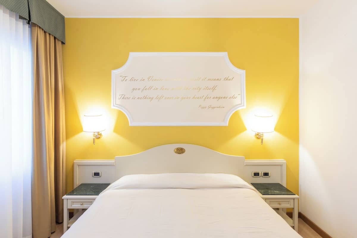 A hotel room with a bed, two bedside tables, a wall-mounted quote, and two wall-mounted lamps.